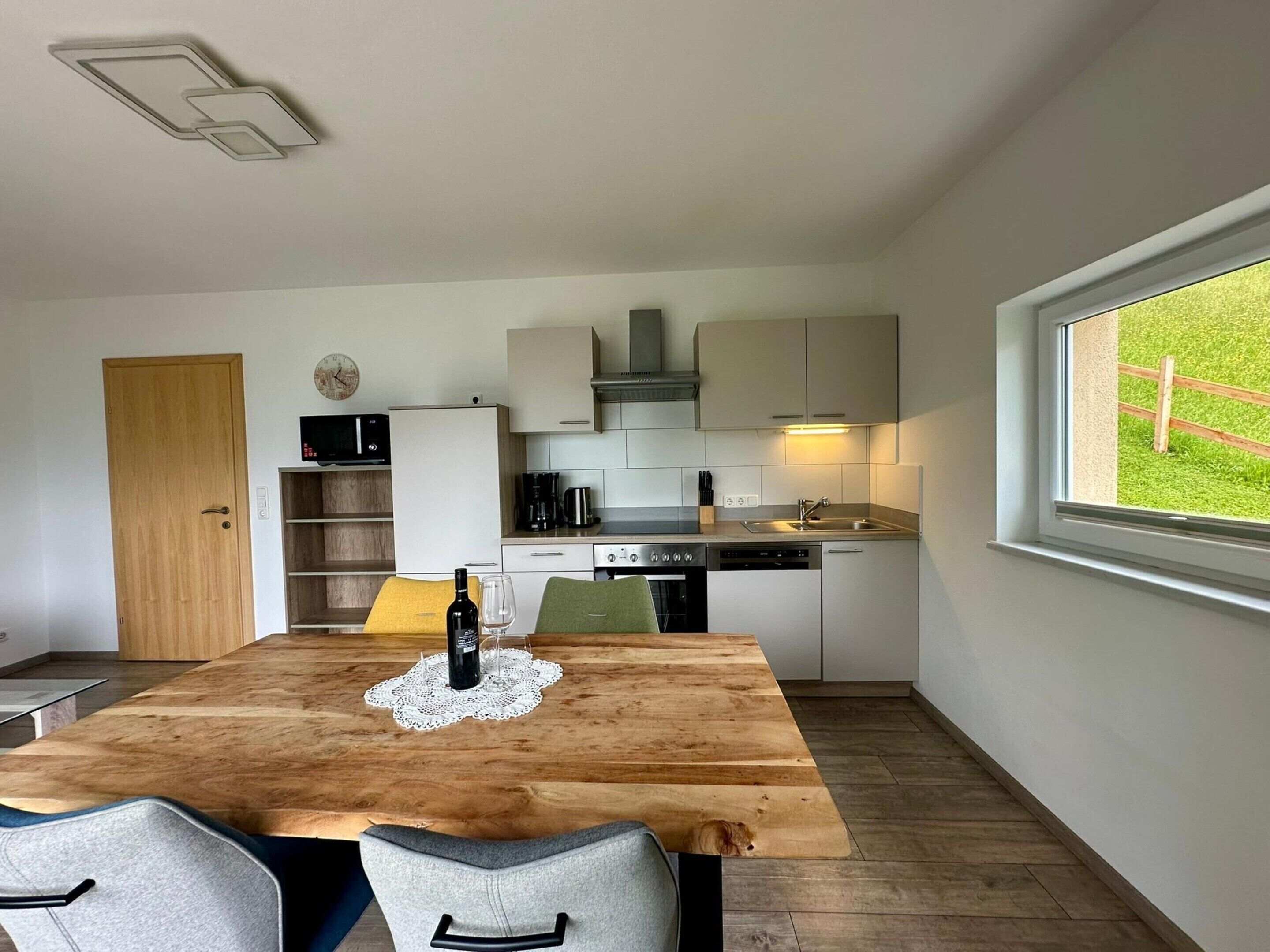 A compact kitchen with modern appliances, a dining table with chairs, and a window with a view of a green landscape.