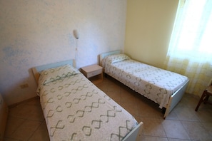 A room with two single beds, a small bedside table, a wall socket, and a window with a curtain. - Beachside Holiday Home in Ricadi (Ricadi)