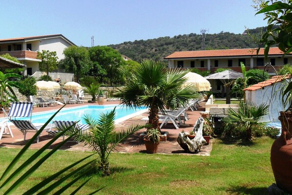 Zona de piscina con sillas de descanso y sombrillas, rodeada de vegetación y edificios con tejados rojos.
