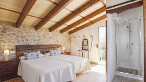 Une chambre avec deux lits, un mur en pierre, des poutres en bois, une table de chevet, une commode, un miroir et une douche fermée par une paroi de verre.