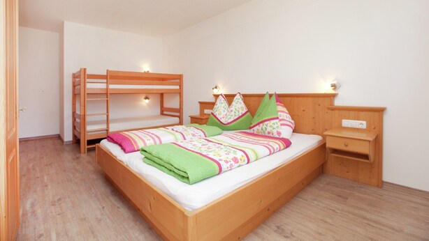 Une chambre avec un lit superposé, un lit simple avec une couvre-lit colorée, une table de chevet et une étagère fixée au mur.