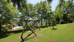 Un'area verdeggiante con un'altalena di legno, circondata da alberi e una vista lontana di un edificio.