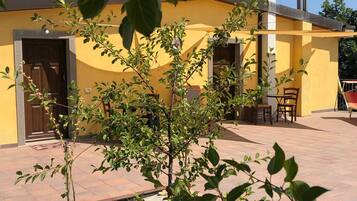 Ein gelbes Gebäude mit Tür und Fenstern, ein kleiner Tisch mit Stühlen und ein Baum im Vordergrund.