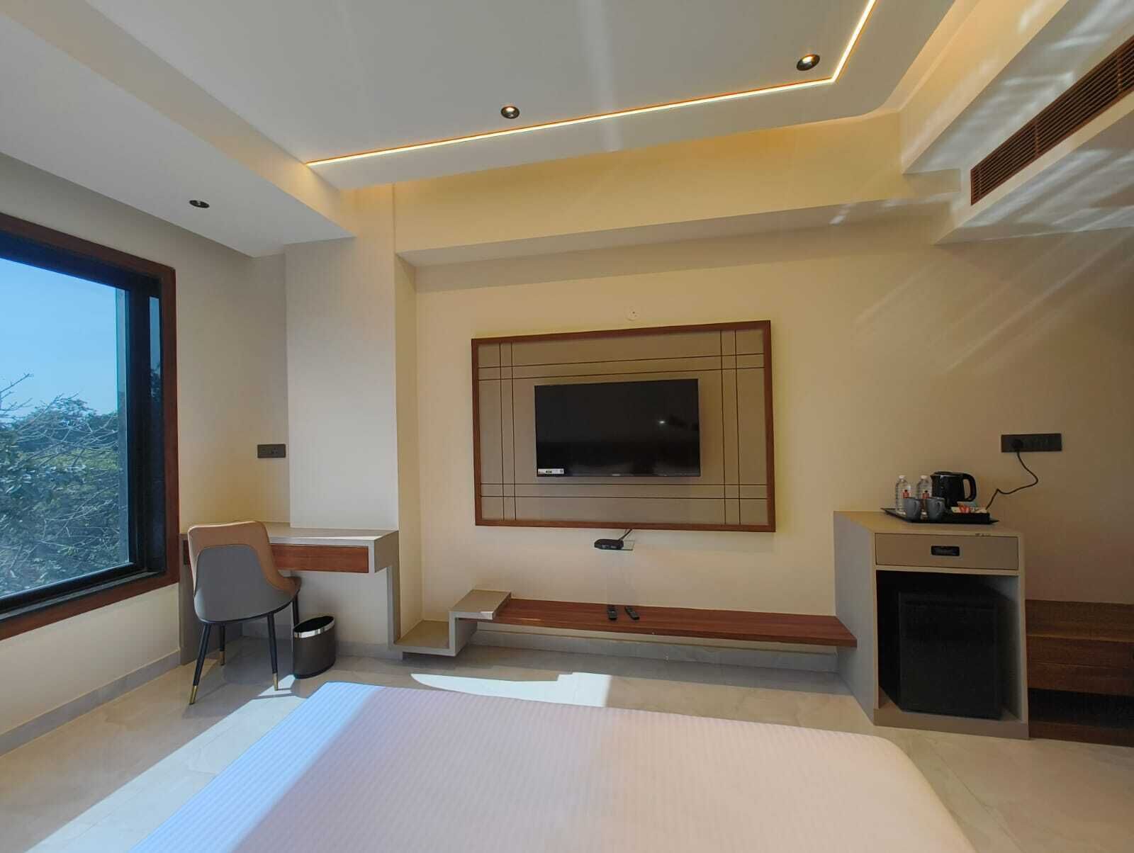 A modern hotel room with a large window, a desk with a chair, a mounted TV, and a small seating area.