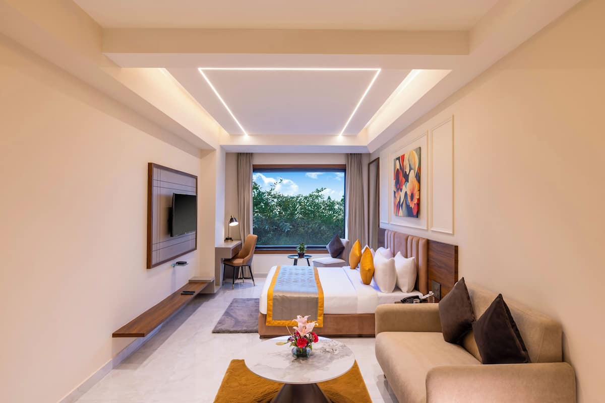 A modern hotel room with a flat-screen TV, a sofa, a coffee table with a vase, a bed with a headboard, a desk with a lamp, and a large window with a view of trees.