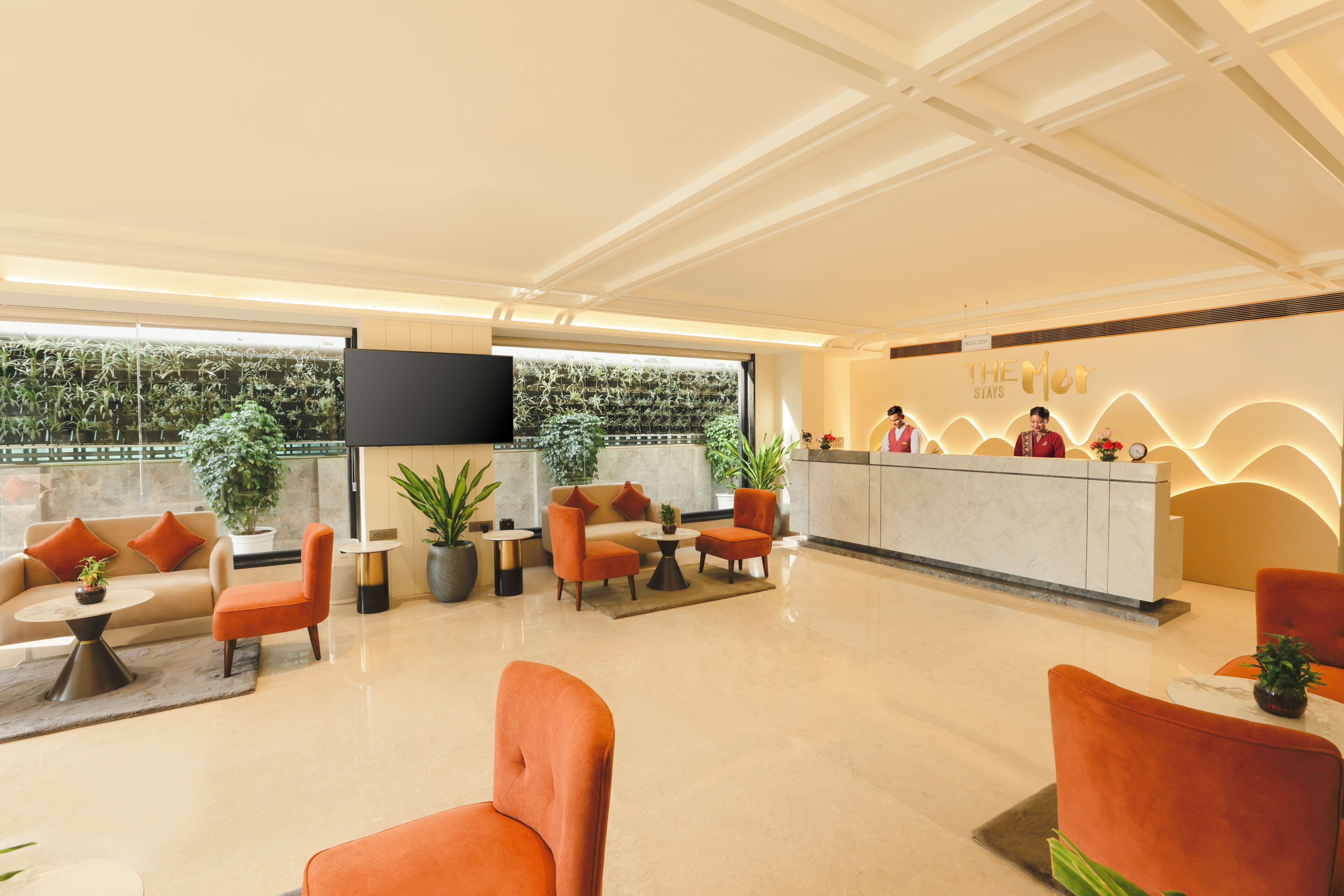 A hotel lobby with a reception desk, seating area, and large windows with plants.