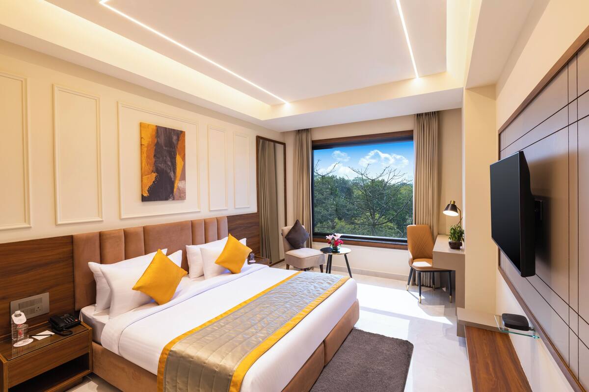 A hotel room with a large bed, a desk, a TV, and a window with a view of trees.
