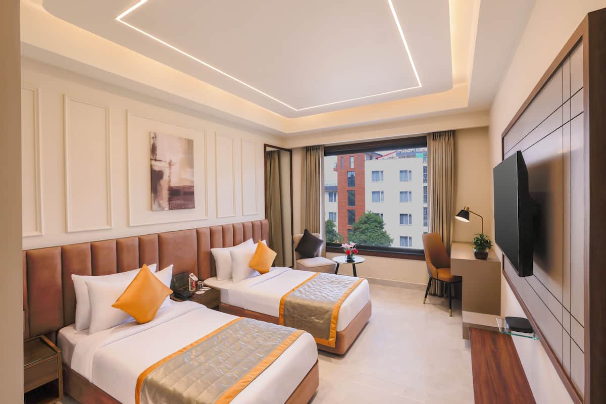 A hotel room with two beds, a desk, a TV, and a view of buildings.