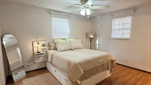 Quarto com cama, ventoinha no teto, mesa de cabeceira, espelho e duas janelas com persianas.