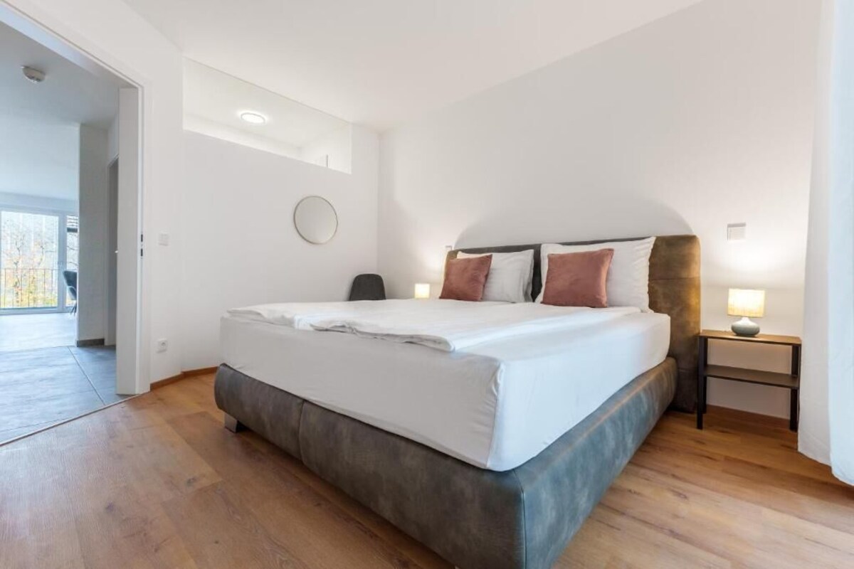 A modern bedroom with a large bed, a bedside table with a lamp, and a view of the outdoors through a door.