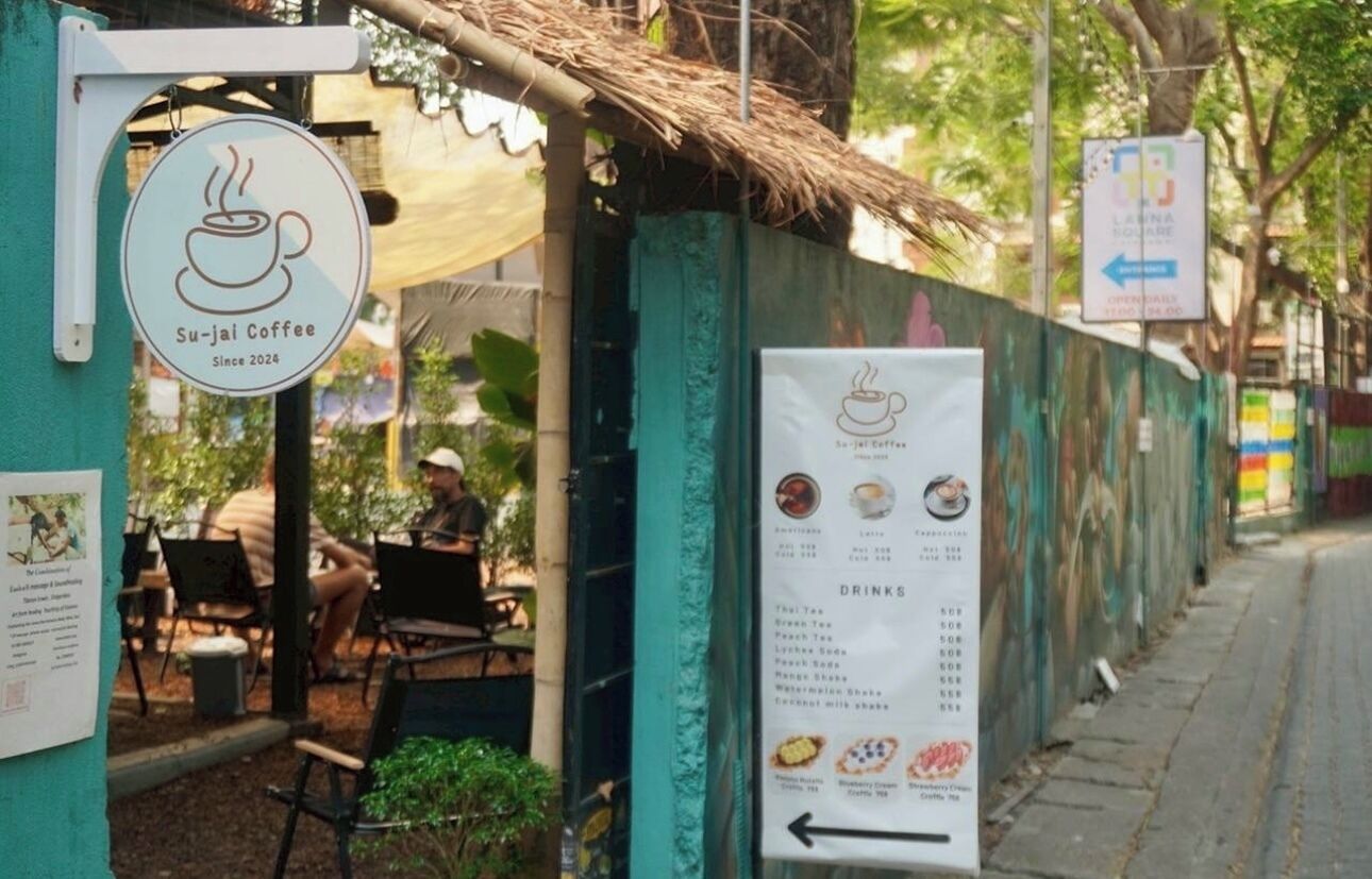 Letrero de Su-jai Coffee y un cartel de menú frente a un edificio de paredes verdes y techo de paja.