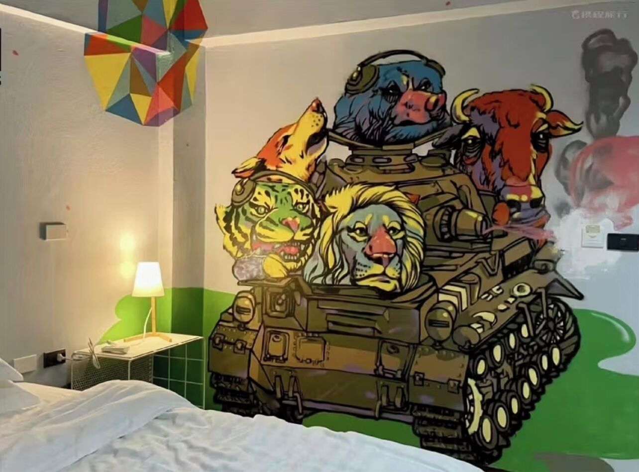 A room with a bed, a lamp, and a wall mural featuring a tank with animal heads.