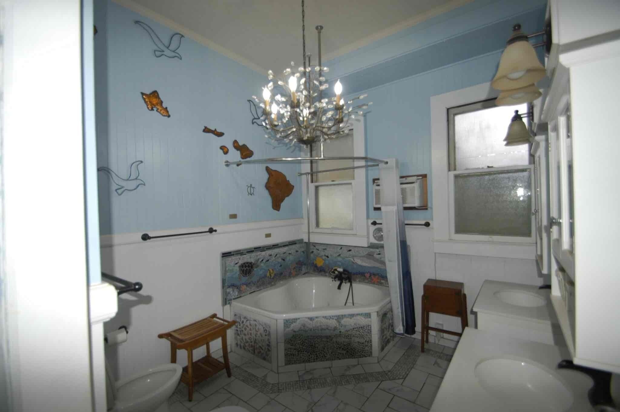 Un baño con bañera, dos inodoros, un banco de madera, una araña y azulejos decorativos.