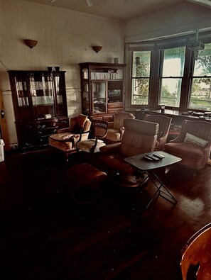 A spacious living room with a large wooden cabinet, multiple chairs, a small table, and a view through large windows. - 3 Suites BnB 2 persons per suite 1 night min  breakfast inc. (Naalehu)