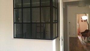Ein Zimmer mit großem Fenster, einer Tür zu einem anderen Raum und einem Sessel in der Ecke.