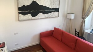Ein Zimmer mit einem roten Sofa, einem Gemälde von einer Berglandschaft, einer Lampe und einem fenster mit Vorhängen.