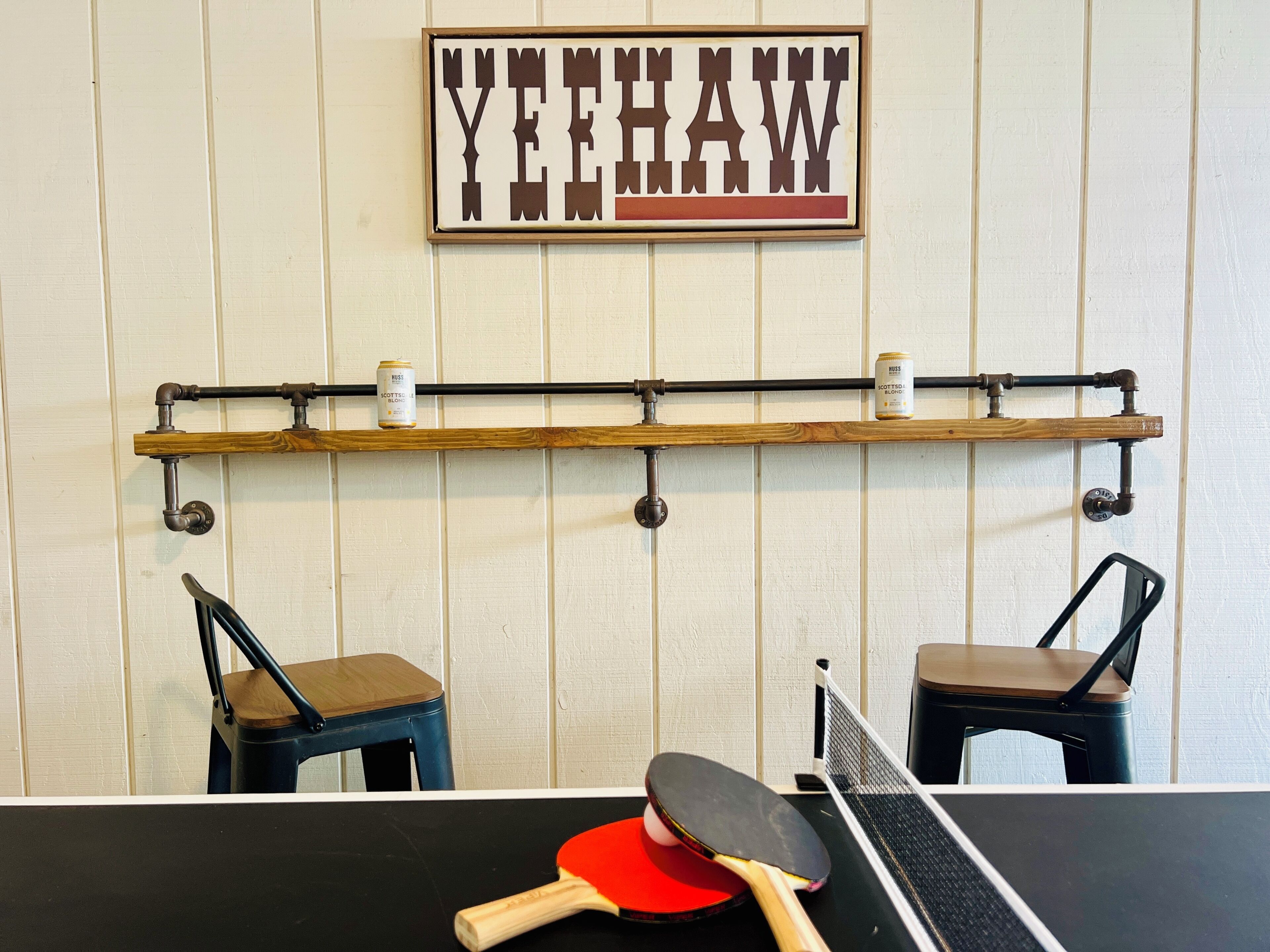 A ping pong table with a table tennis paddle and two chairs, a shelf with cans, and a wall sign.
