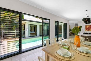 A modern dining area with a large glass door leading to a pool, a dining table set with plates and glasses, and a decorative orange vase. - Belle Escapes Sand Castaways (Palm Cove)