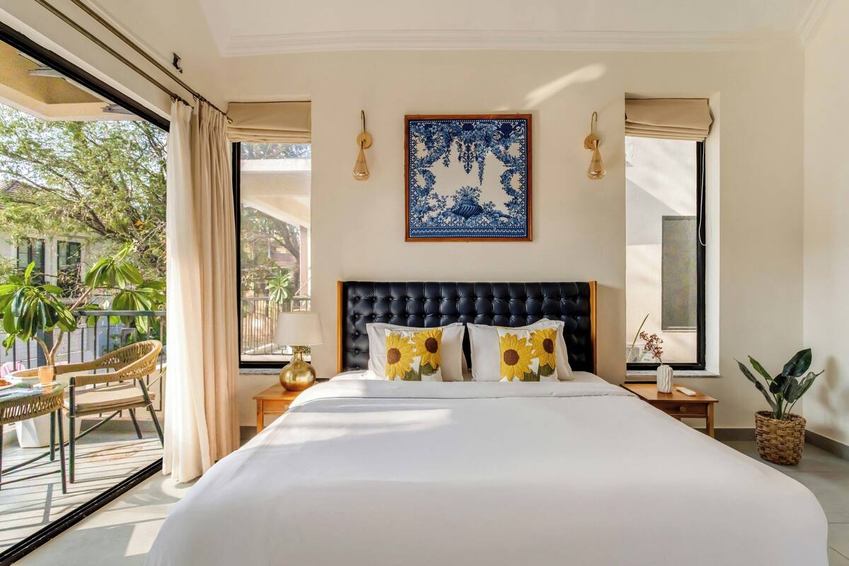 A hotel room with a large bed, a view of the outdoors, a decorative wall piece, and a small table with a lamp.