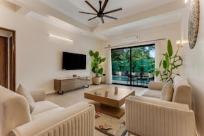 A modern living room with a flat-screen TV, a ceiling fan, and a large sliding glass door leading to an outdoor area with plants.
