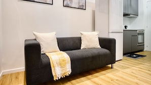 Un salon moderne avec un canapé gris foncé, deux coussins blancs et un plaid à carreaux jaunes et blancs. On y trouve également un coin cuisine avec des meubles gris et des appareils électroménagers blancs.