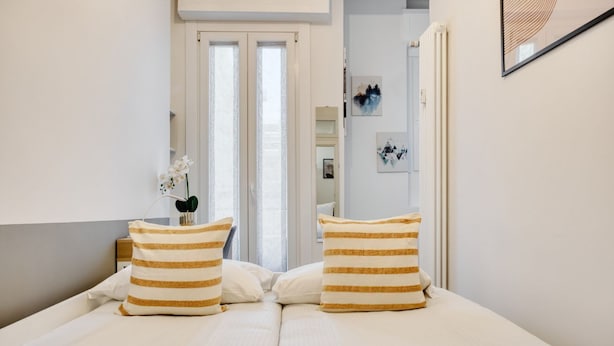 Une chambre à coucher avec un lit, des coussins à rayures, une table de chevet, un vase avec des fleurs et une porte donnant sur une autre pièce.