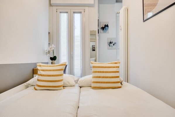 Un dormitorio con cama, almohadillas a rayas, una mesita de noche, un florero con flores y una puerta que da a otra habitación.