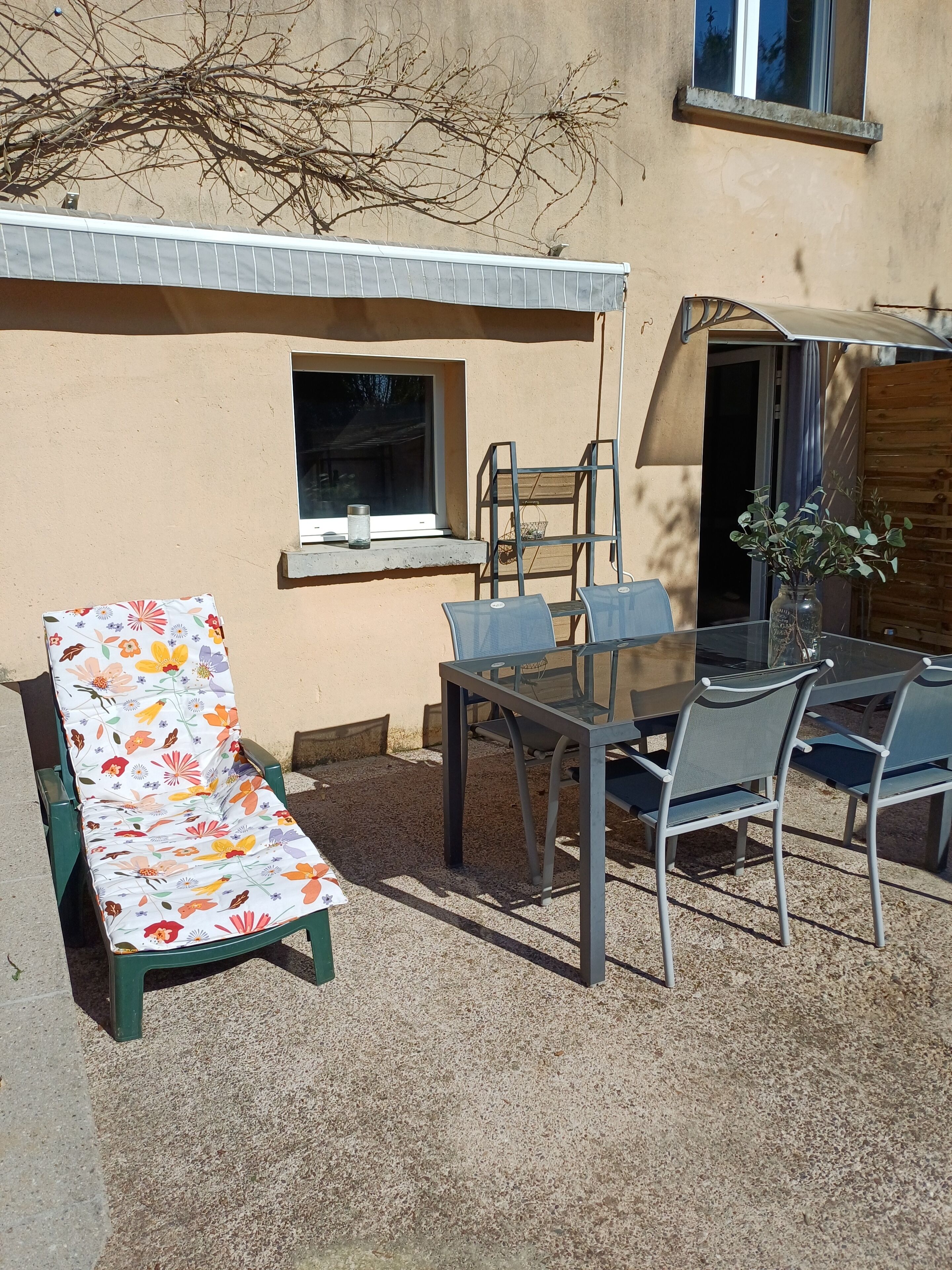 Een terras met een tafel en stoelen, een bank met een bloemenkussen, en een raam met uitzicht op de lucht.