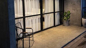 A modern building exterior with large glass windows, a concrete facade, and a wooden deck area with a chair and potted plants.