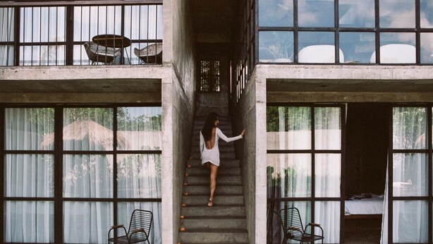 A woman descending a concrete staircase in a building with large windows and two chairs at the bottom.