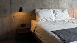 A bedroom with a concrete wall, a bed with white linens, a black throw blanket, and a pendant light.