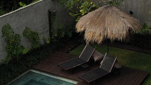 A poolside area with a wooden deck, two lounge chairs, a thatched umbrella, and a small table.