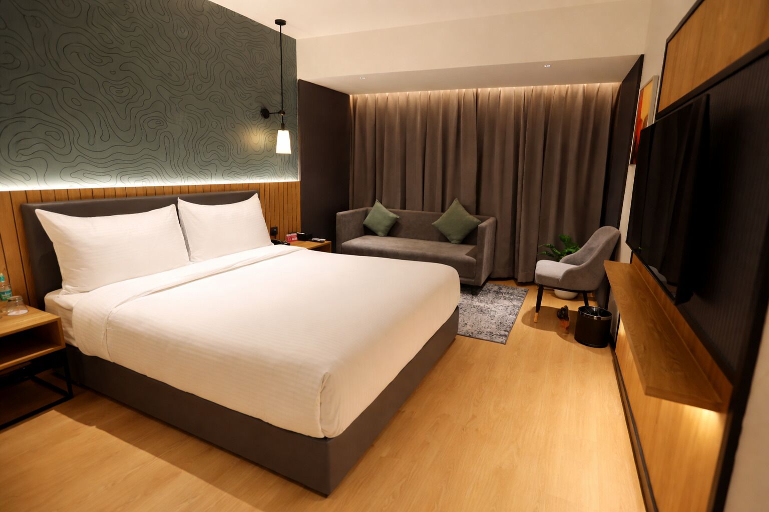 Une chambre d’hôtel moderne avec un grand lit, un canapé, un fauteuil, une télévision et un plancher en bois.