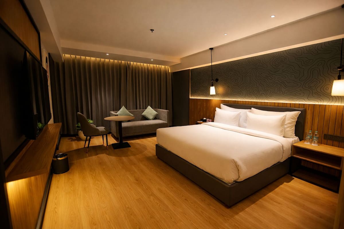 A modern hotel room with a large bed, a desk, a chair, a TV, and a wall-mounted lamp.