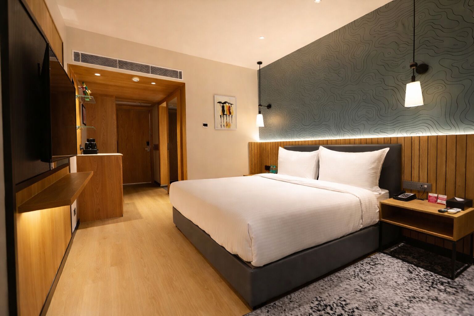 A modern hotel room with a large bed, wooden headboard, bedside tables, a flat-screen TV, and a unique wall design.