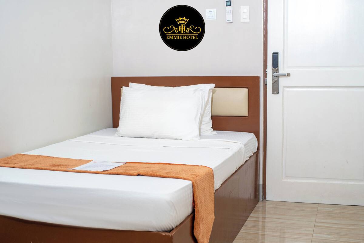A single bed with a wooden headboard, white bedding, a brown throw blanket, and a wall-mounted emblem of 'EMMIE HOTEL'.