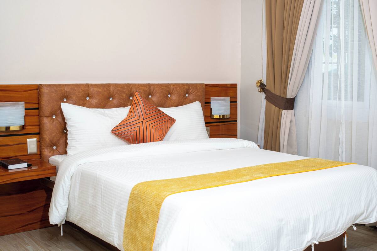 A neatly made bed with a headboard, a bedside table with a lamp, and a window with curtains.