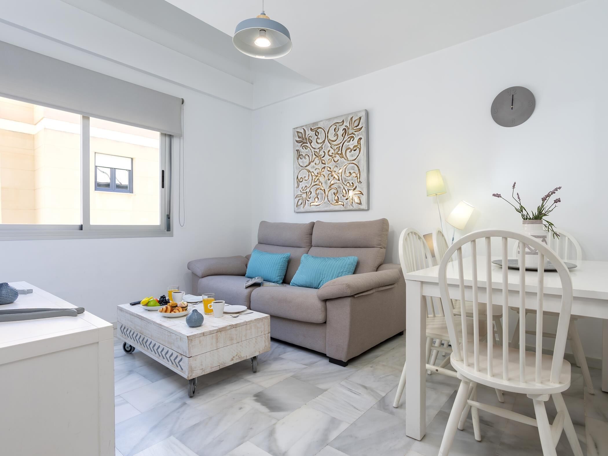 A modern living room with a sofa, a coffee table, a dining table with chairs, a wall clock, and a window.