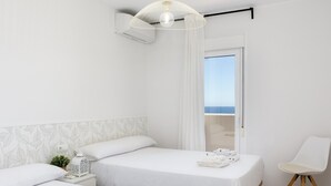 Una habitación de hotel moderna con dos camas, una mesita de noche con lámpara, una silla y vista al mar.