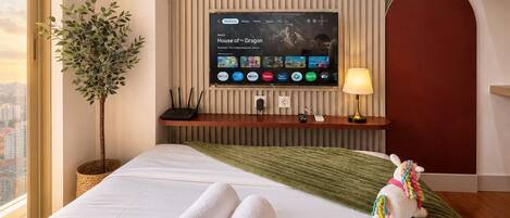 Une chambre d’hôtel avec un lit, un téléviseur fixé au mur, une plante et un jouet en peluche.