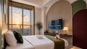 A hotel room with a large bed, a TV, a plant, and a view of the cityscape. - Arlo By SanLiving - 2BR - HubLife Mall (Jakarta)