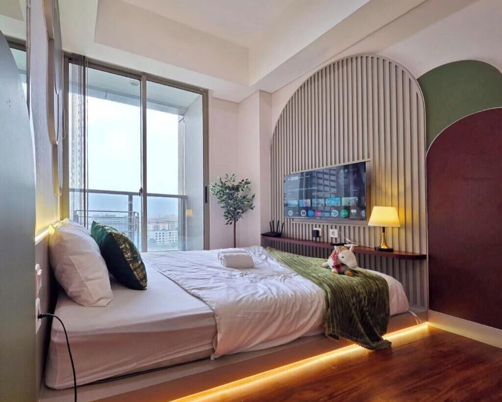 Une chambre d’hôtel moderne avec un grand lit, un téléviseur fixé au mur, une fenêtre donnant sur la ville et une plante posée sur le rebord de la fenêtre.