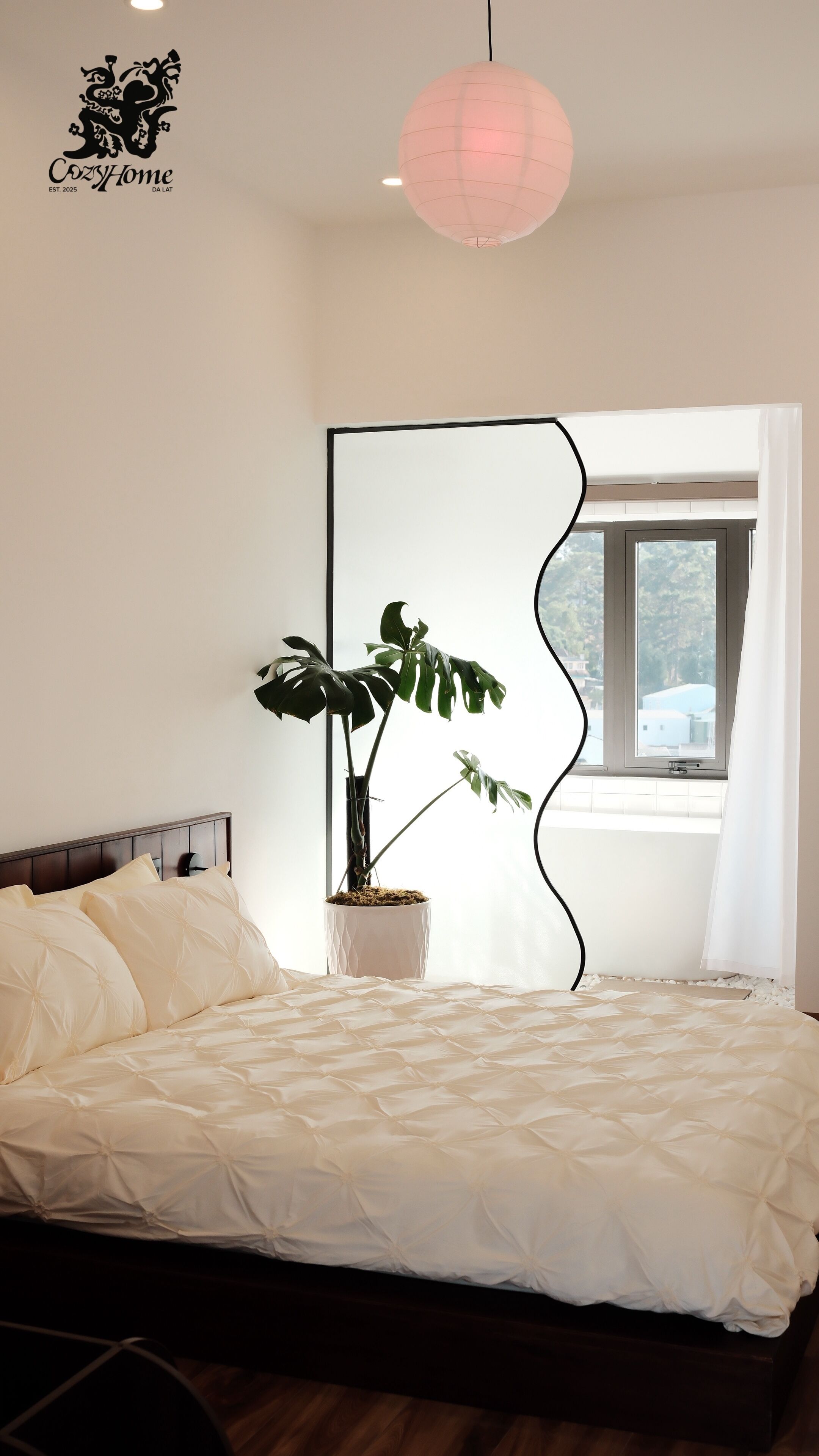 Un dormitorio con una cama, una planta en maceta, una ventana con cortinas y una lámpara fijada en la pared.