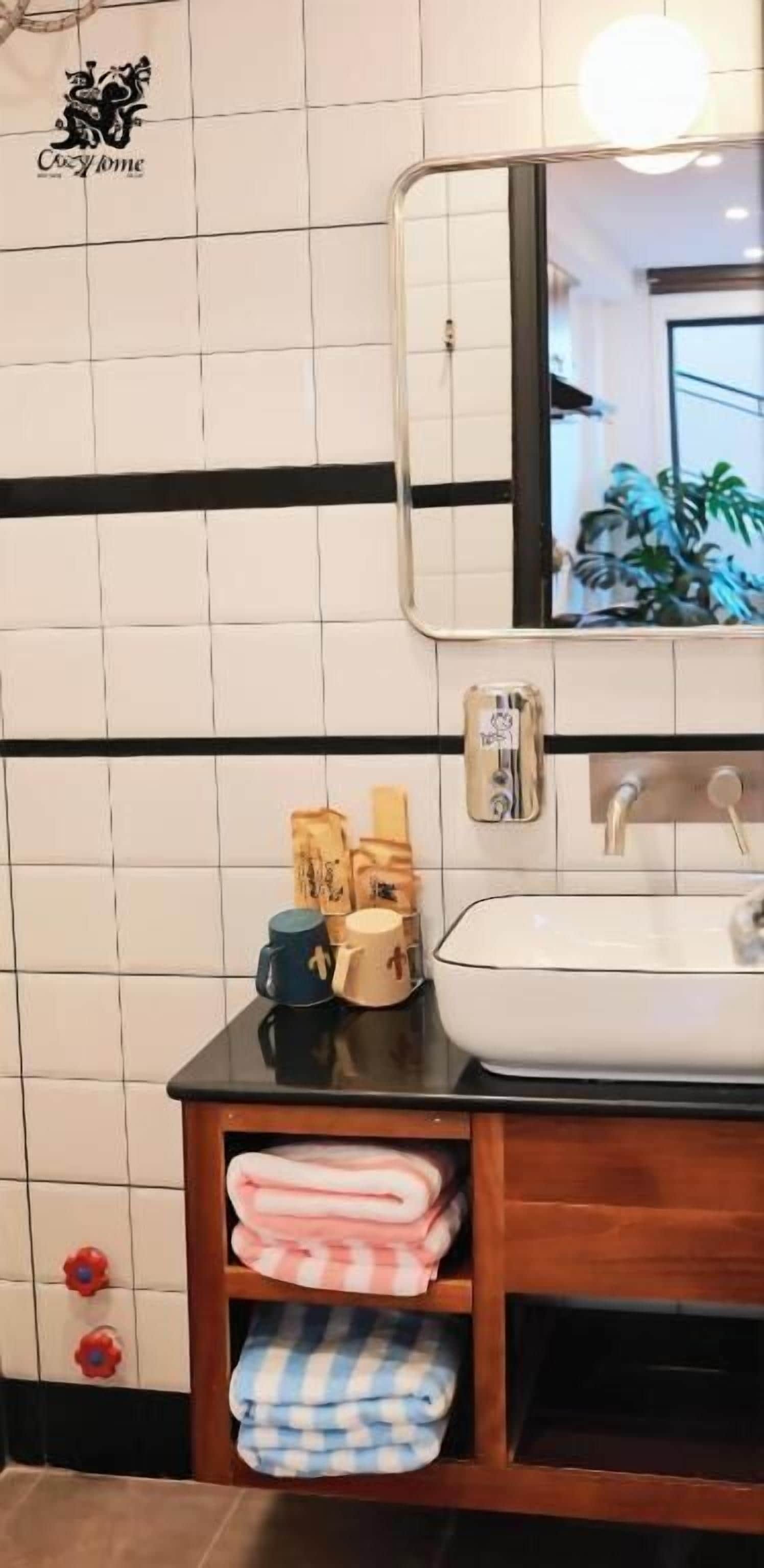 Un baño con paredes de azulejos blancos, encimera negra, un lavabo y un espejo. Hay toallas y un dispensador de jabón en la encimera.