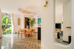 A spacious room with a tiled floor, a wooden dining table, and chairs. There is a large window with curtains, a plant, and a view of greenery outside. - Vida Pacifica Villa (Verla Canca)