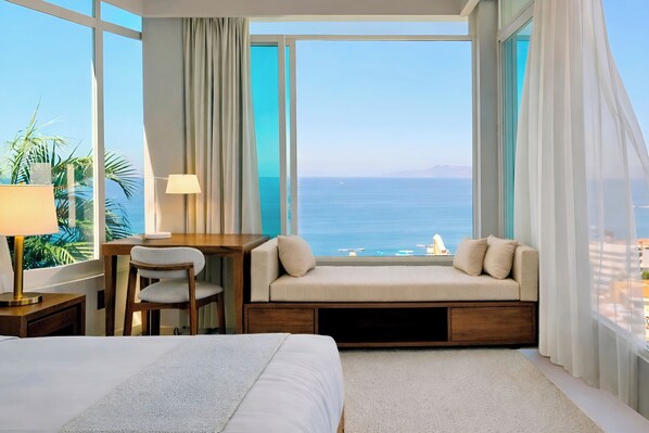 Ein Hotelzimmer mit großem Fenster und Meerblick, einem Bett, einem Schreibtisch mit Stuhl sowie einer kleinen Sitzecke mit Kissen.