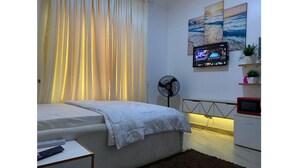A bedroom with a bed, a TV mounted on the wall, a fan, and a small shelf with a plant. - Cozy 1-bedroom house with 247 power, FREE Wifi, Netflix, AC (Lekki)