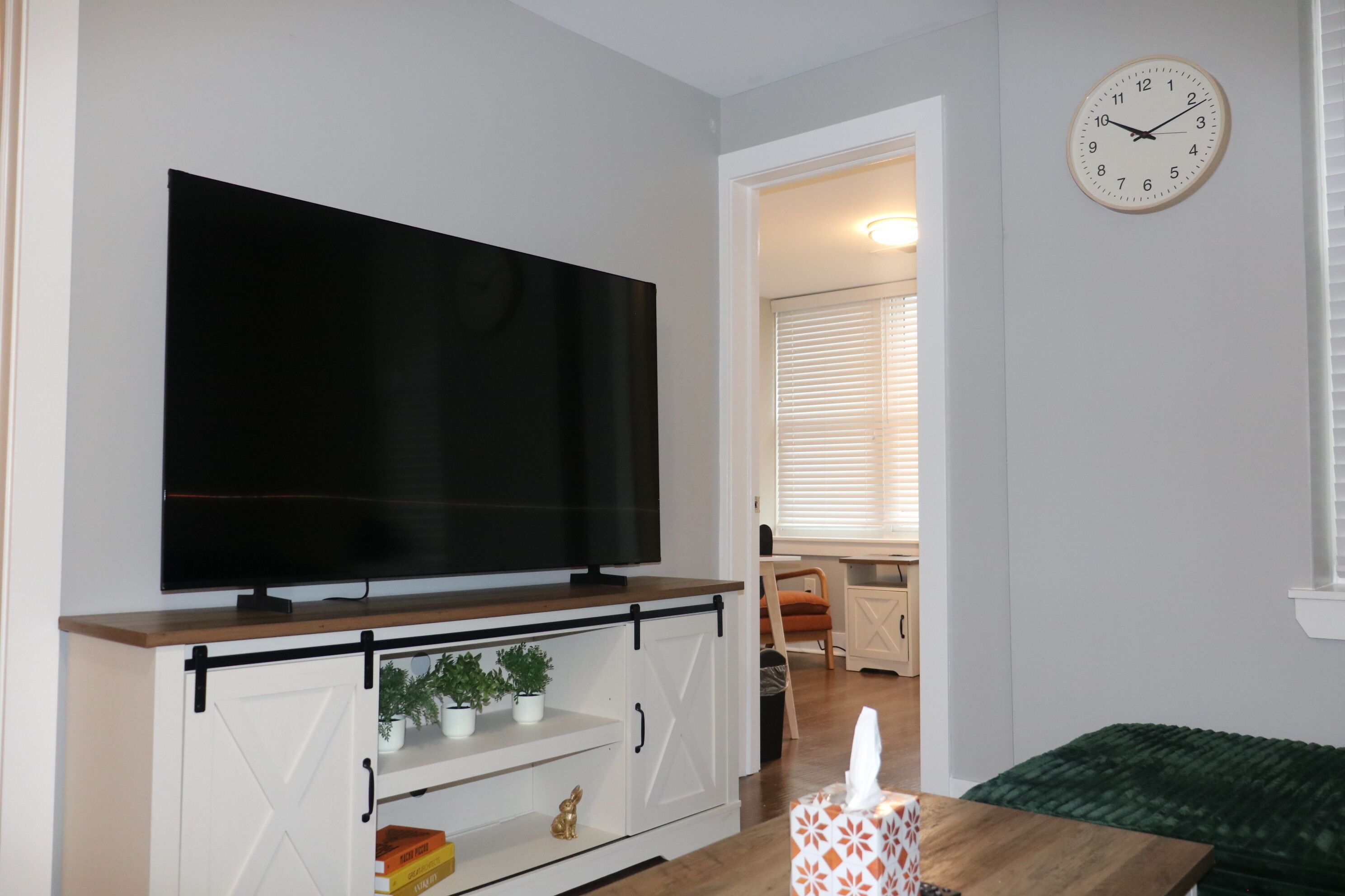 Un salon avec un téléviseur à écran plat posé sur une étagère en bois, un meuble blanc orné d’un motif en forme de croix, un canapé vert et une horloge murale.