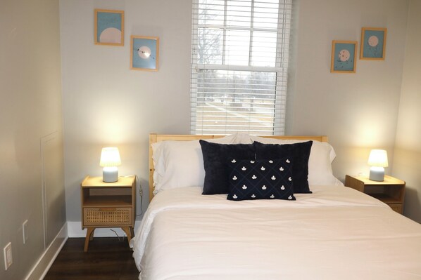 A bedroom with a bed, two bedside tables with lamps, a window with blinds, and wall art.