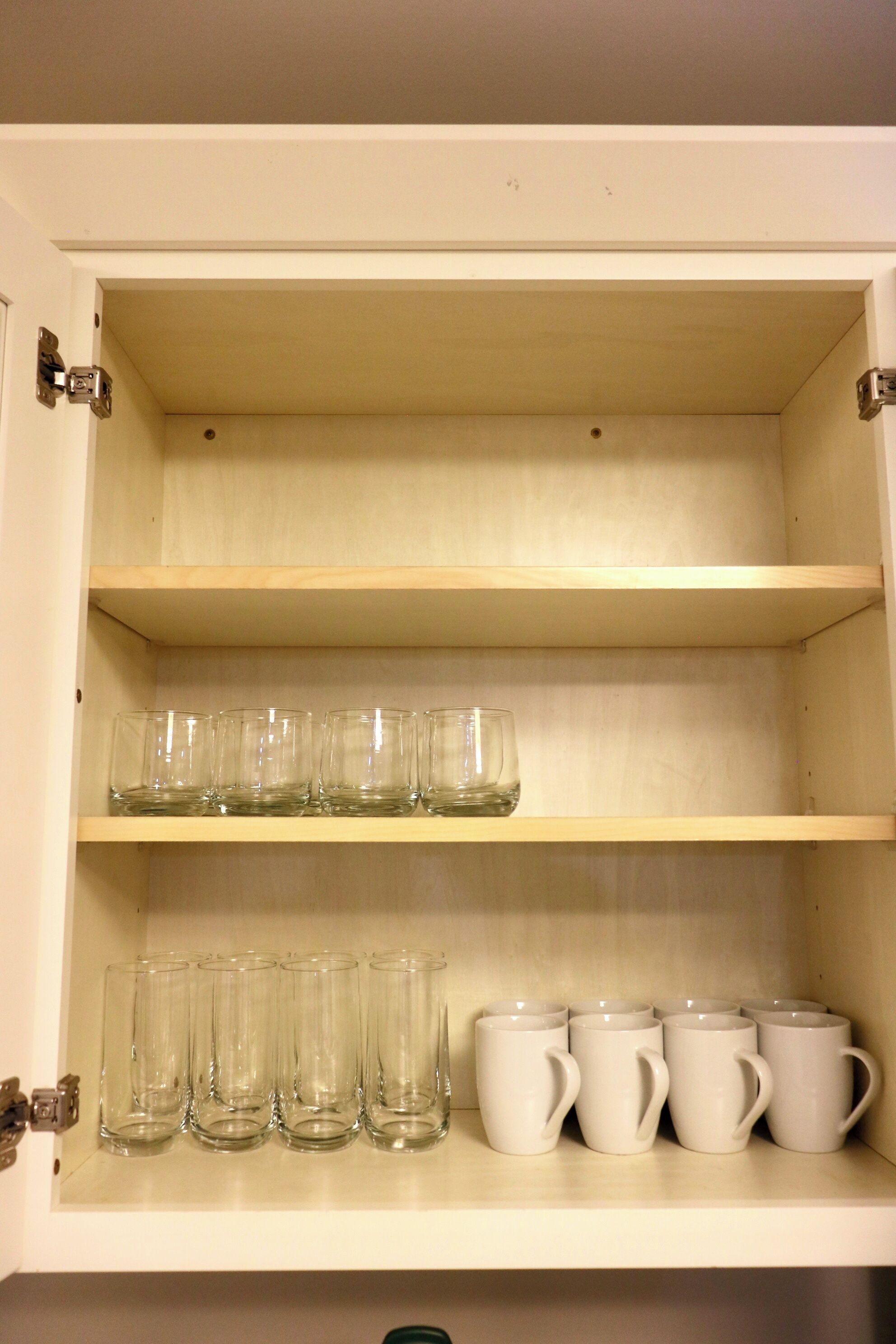 A cabinet with two shelves; the top shelf holds six clear glasses, and the bottom shelf holds six white mugs.